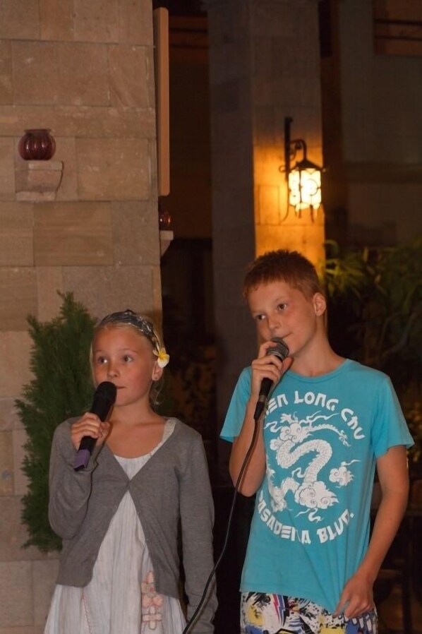 Two individuals holding microphones, standing in front of a stone wall with a decorative item and a wall-mounted light fixture.