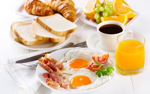 Prato de pequeno-almoço com ovos, bacon e tomate, acompanhado por croissants, torradas, fruta, café e sumo de laranja.