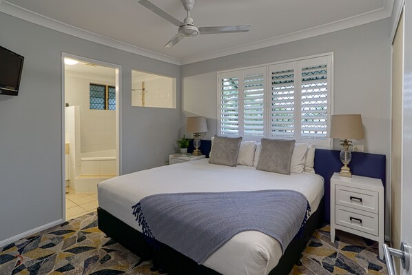 Quarto com cama, duas mesas de cabeceira, ventilador de teto, janela com persianas e porta para um banheiro.