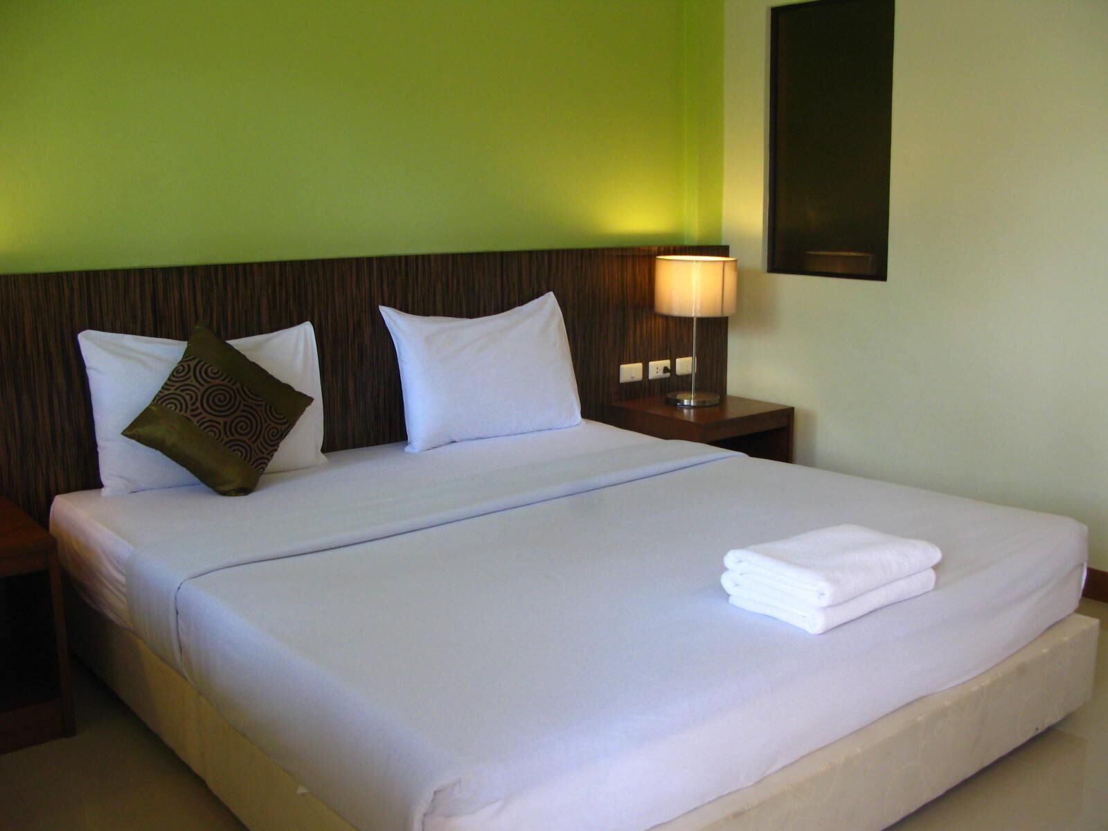 A neatly made bed with white linens and a folded towel, a wooden headboard, a nightstand with a lamp, and a green wall.