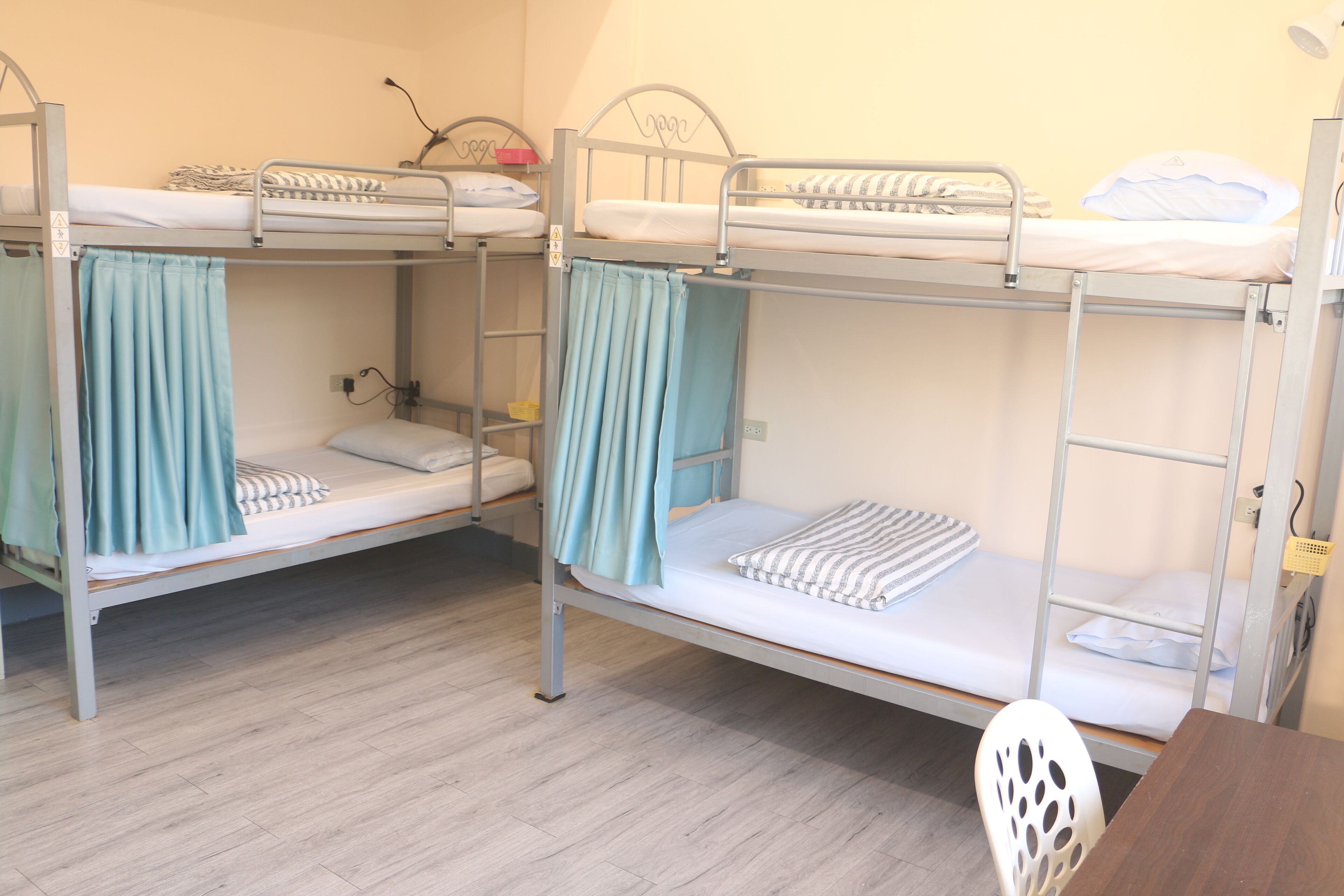 A room with three bunk beds, each with a curtain and a pillow. There is a wooden table and a white chair in the room.