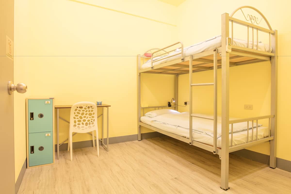 A bunk bed room with a desk and a chair, a small cabinet, and a wall socket.