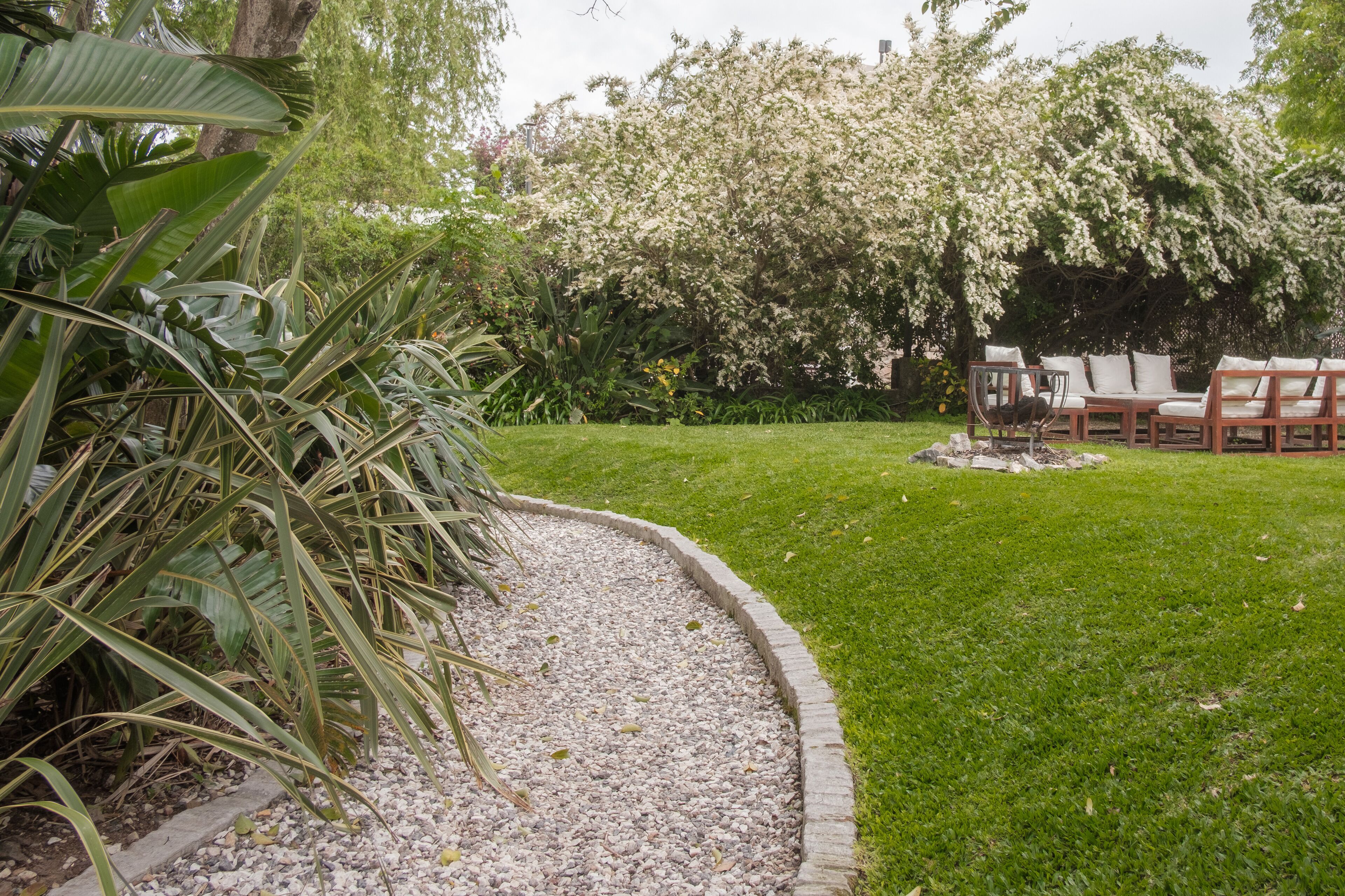 Un jardín con un sendero de grava, un banco de madera y una mesa con sillas.