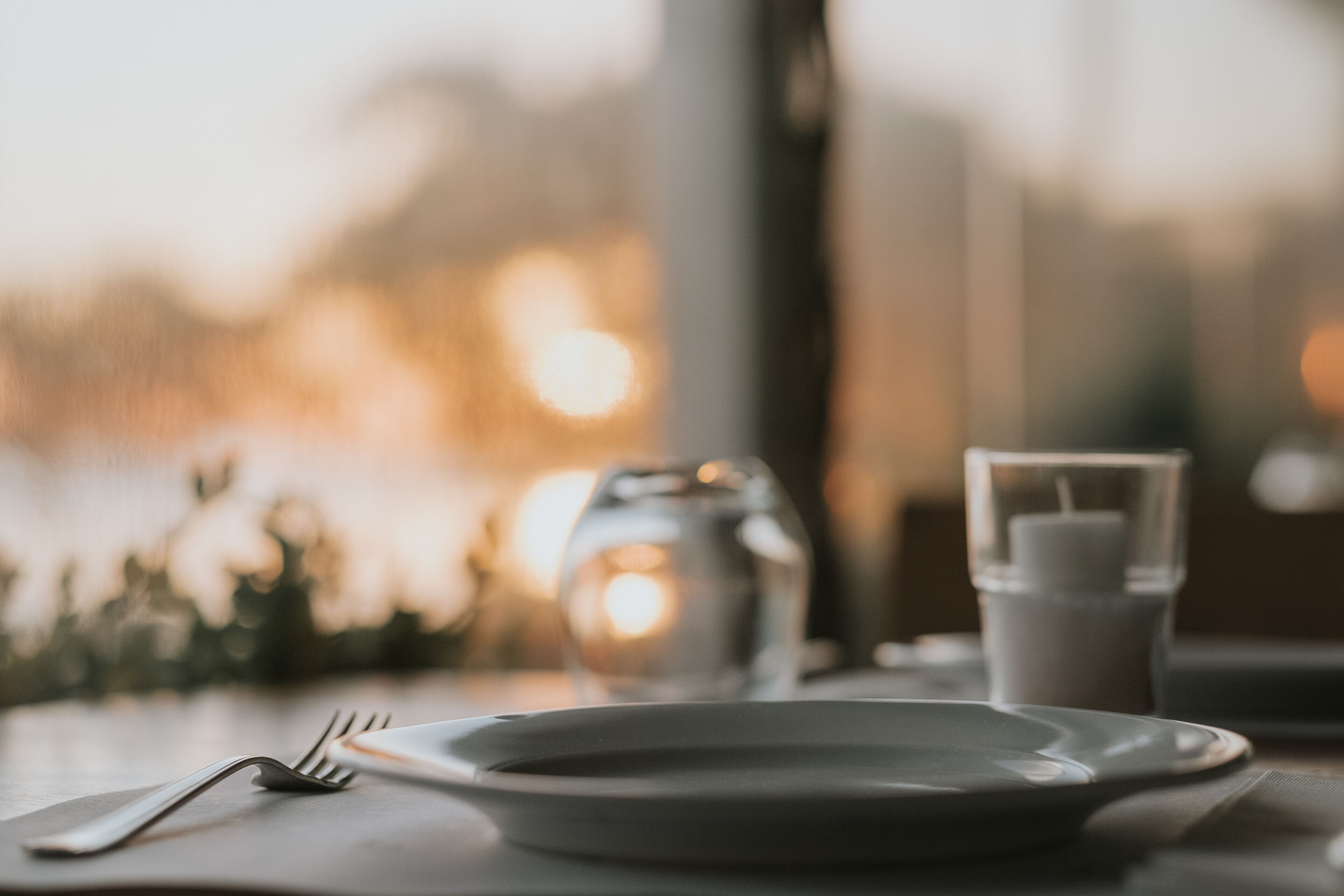 A dining table with a plate, fork, and glass, set against a blurred background with a candle and a vase.