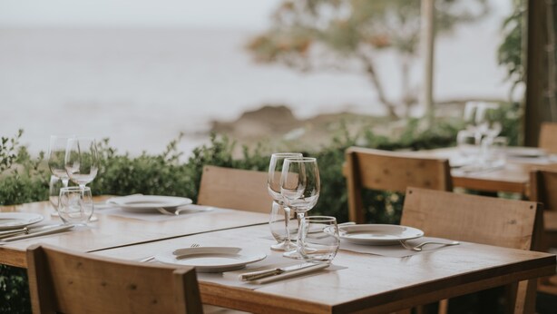 A wooden dining table set with plates, glasses, and cutlery, overlooking a body of water. - Charco Hotel (Colonia del Sacramento)