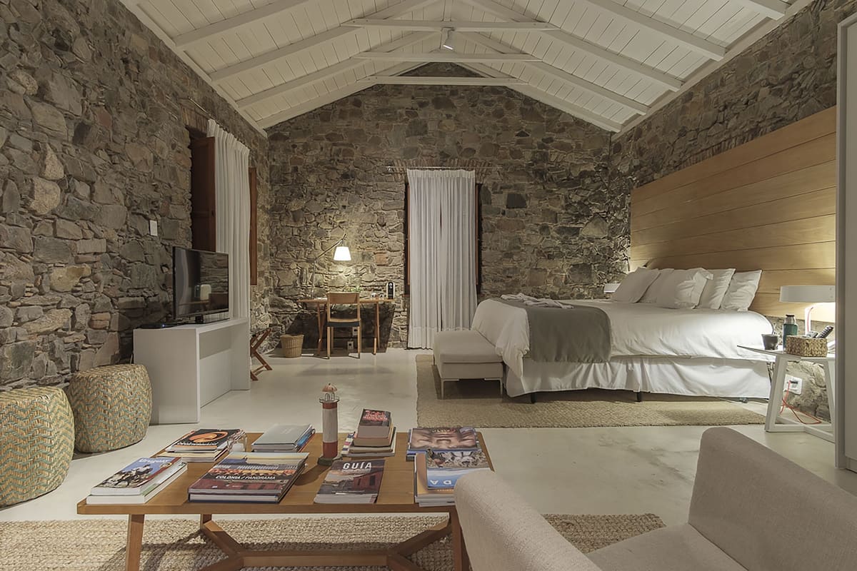 Une chambre confortable avec un mur en pierre, un lit avec une literie blanche, une tête de lit en bois, une table de chevet avec une lampe et un téléviseur posé sur un meuble.