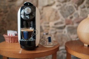 Una cafetera sobre una mesa de madera, con una taza de café y una cesta al fondo.