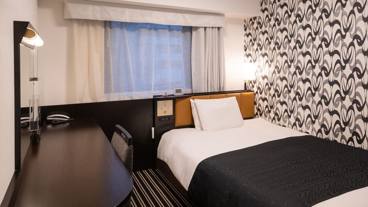 Een hotelkamer met een bed, een bureau, een stoel, een raam met gordijnen en een behang met een patroon.