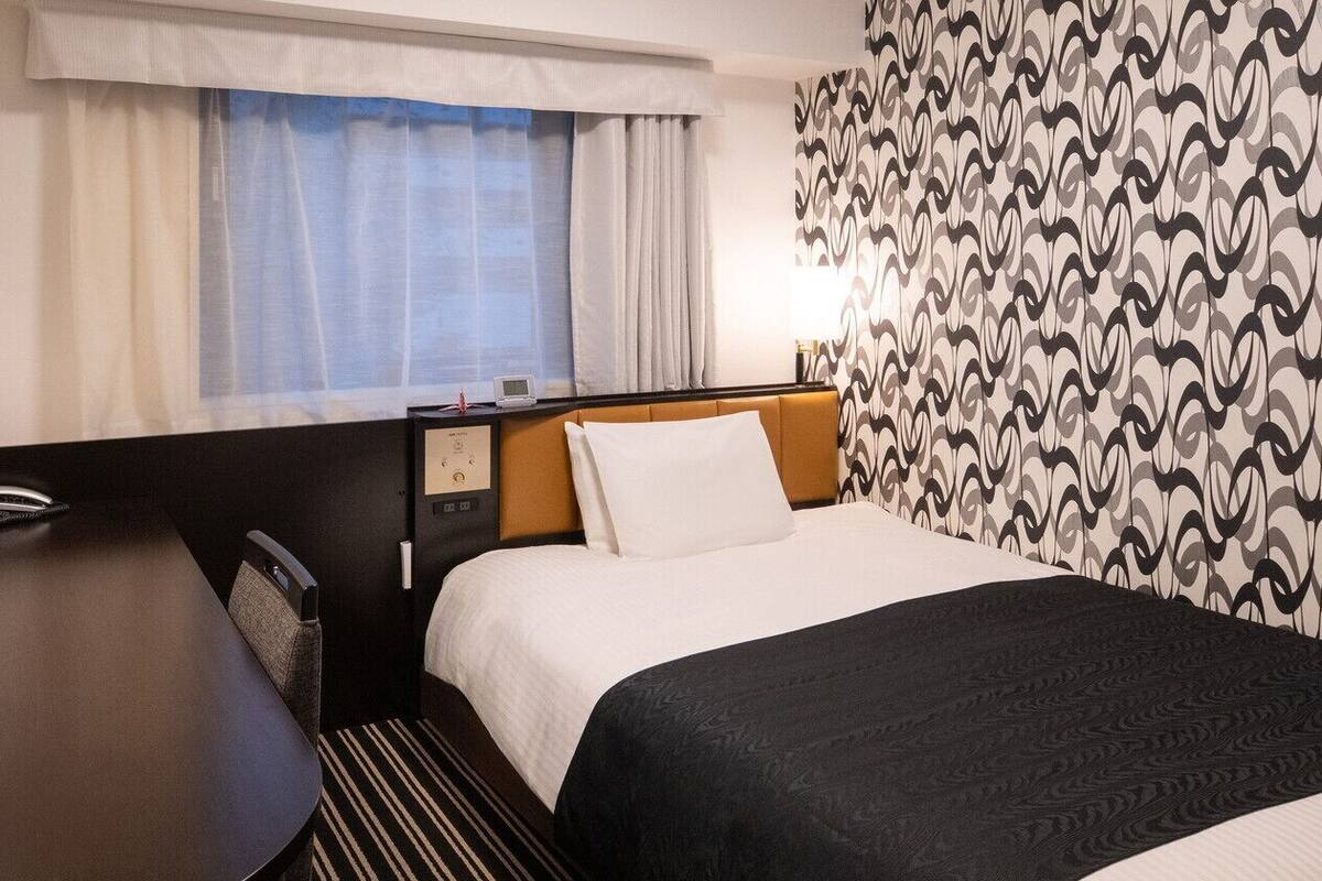 Habitació d'hotel amb un llit, una taula, una cadira, una finestra amb cortines i una paret amb un disseny.