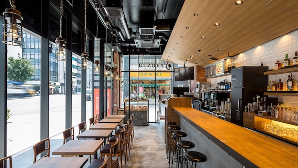 A modern cafe with wooden tables and bar stools, large windows, and a visible street view. - APA Hotel Higashi Nihombashi Ekimae (Tokyo)