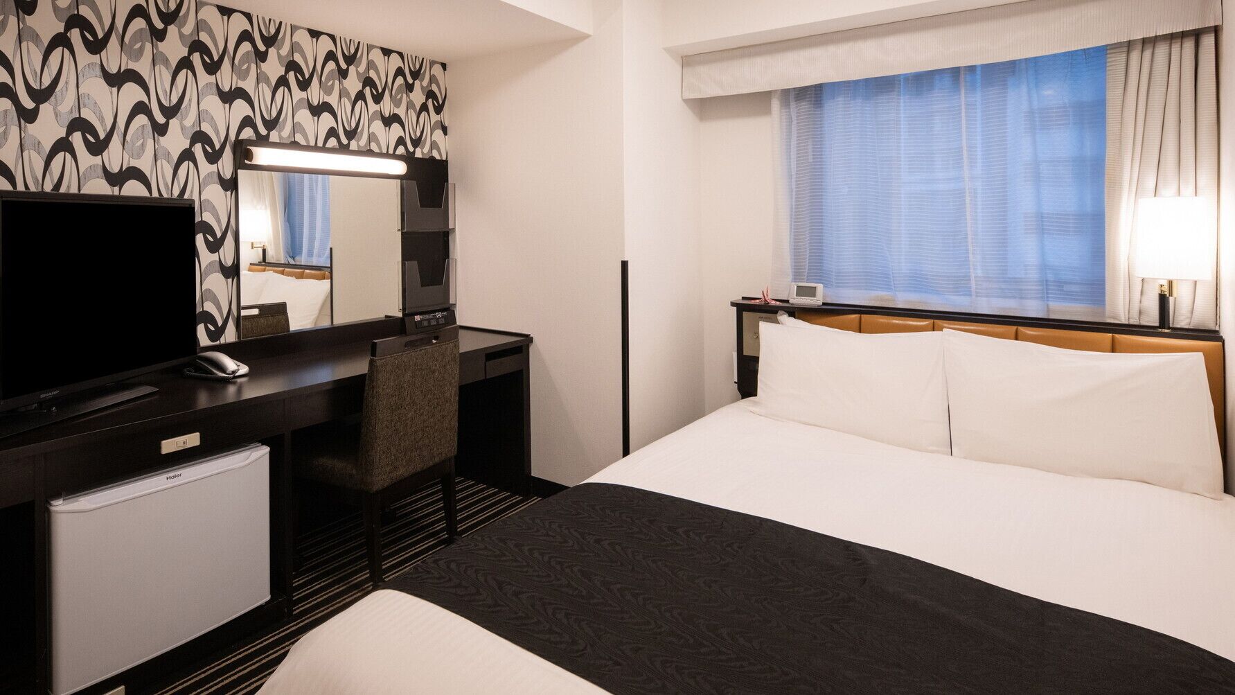 Een hotelkamer met een bed, een bureau met een computer, een stoel, een televisie en een raam met gordijnen.