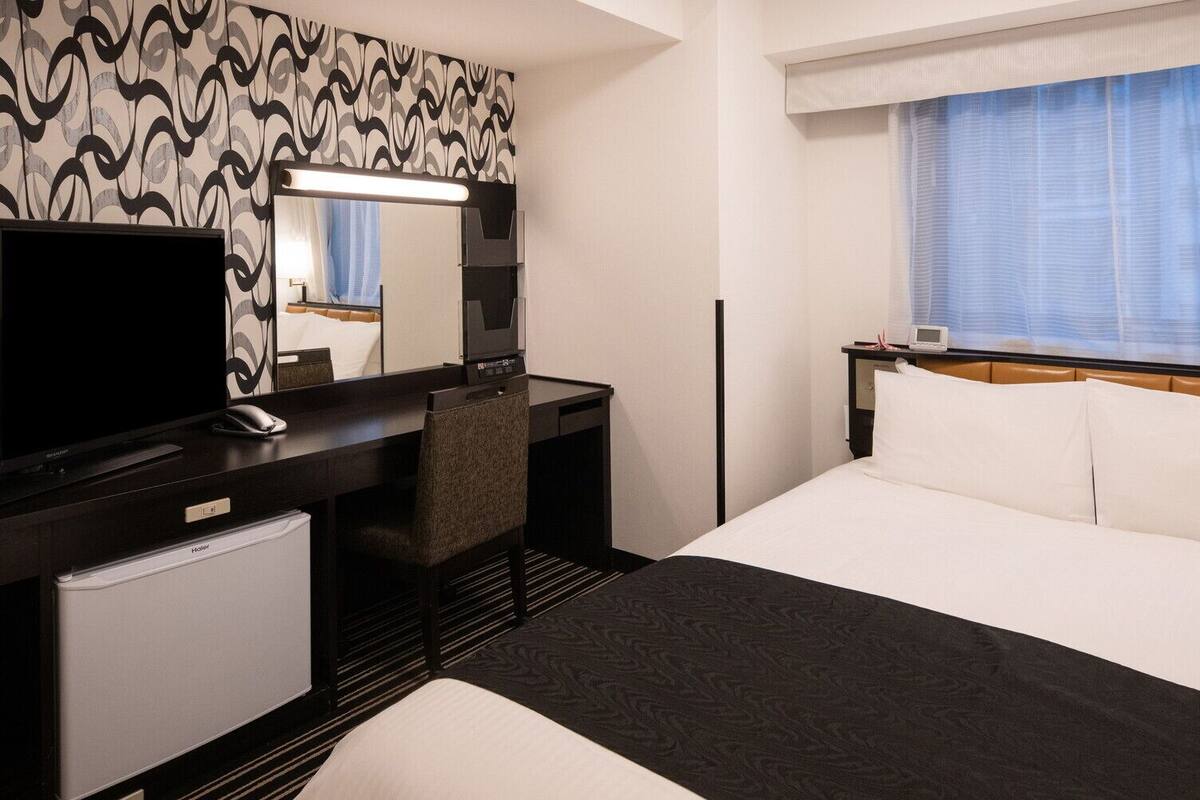 Habitació d'hotel amb un llit, una taula de treball amb un ordinador, una cadira, una televisió i una finestra amb cortines.