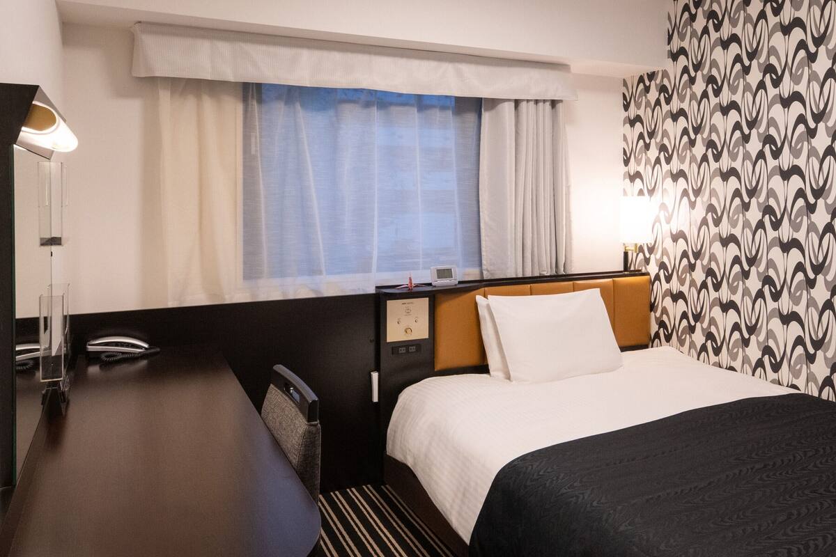 Habitació d'hotel amb llit, escriptori, cadira, tauleta de nit, làmpada i finestra amb cortines.