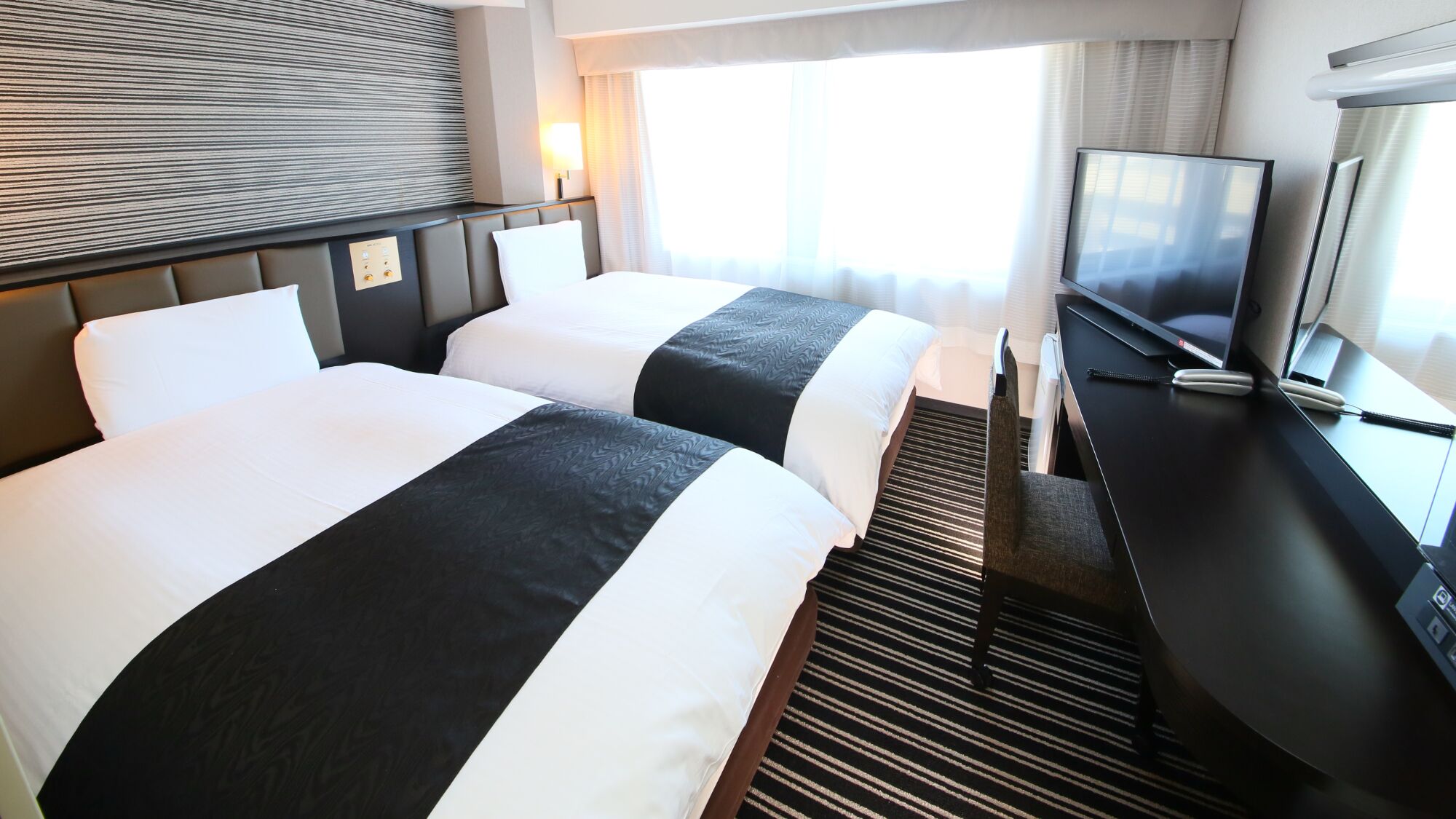 Camera d'albergo con due letti, televisore, scrivania e finestra con tende.