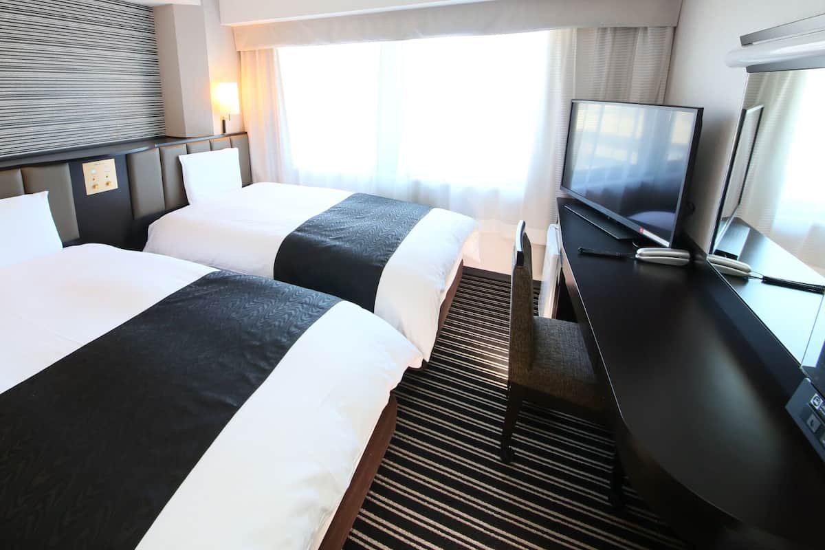 A hotel room with two beds, a TV, a desk, and a window with curtains.