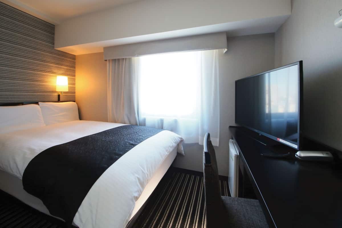 A hotel room with a bed, a flat-screen TV, a desk, and a window with curtains.