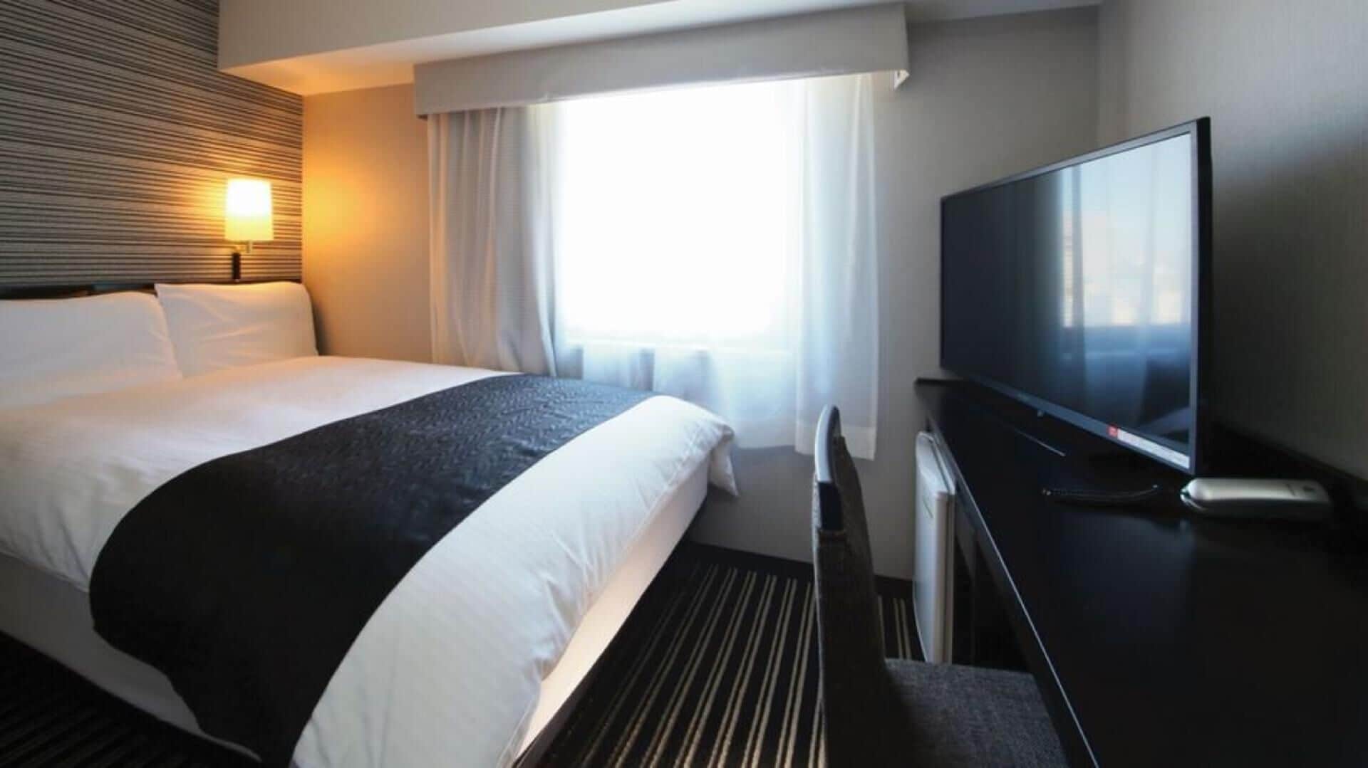 Una camera d'albergo con un letto, una TV, una finestra con tende e una lampada a parete.