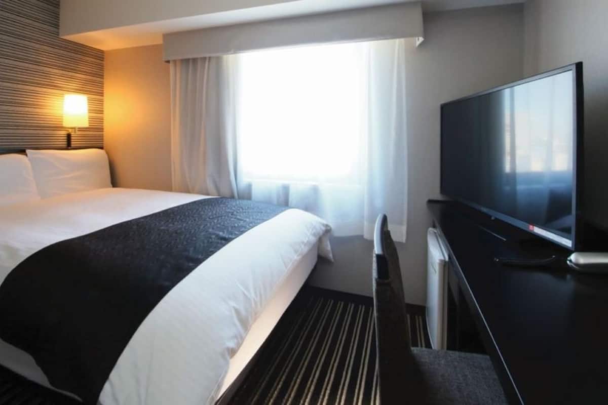 A hotel room with a bed, a TV, a window with curtains, and a wall-mounted light.