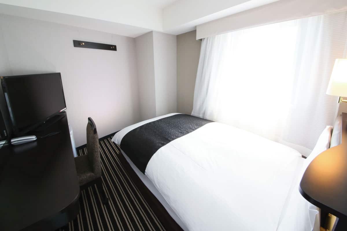A hotel room with a bed, a desk with a television, a chair, a window with curtains, and a bedside table with a lamp.