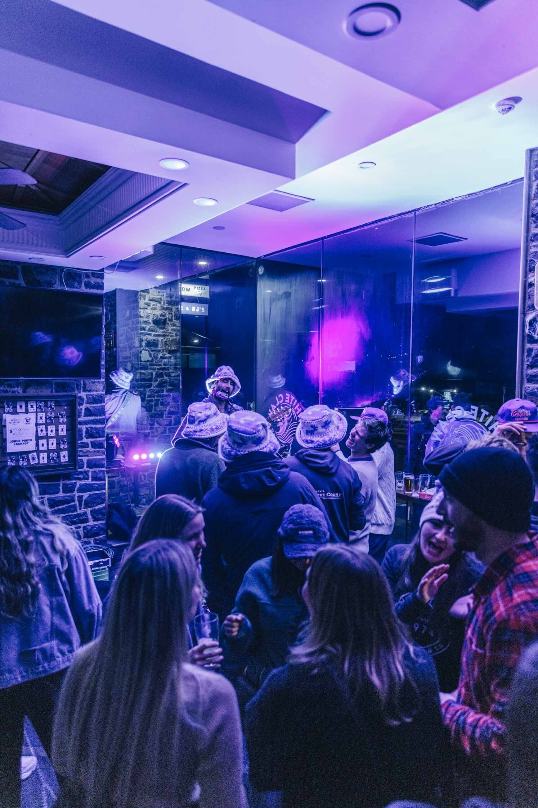 Un bar concurrido con gente usando sombreros y chaquetas, un letrero que dice 'Pizza' y una pared de piedra.