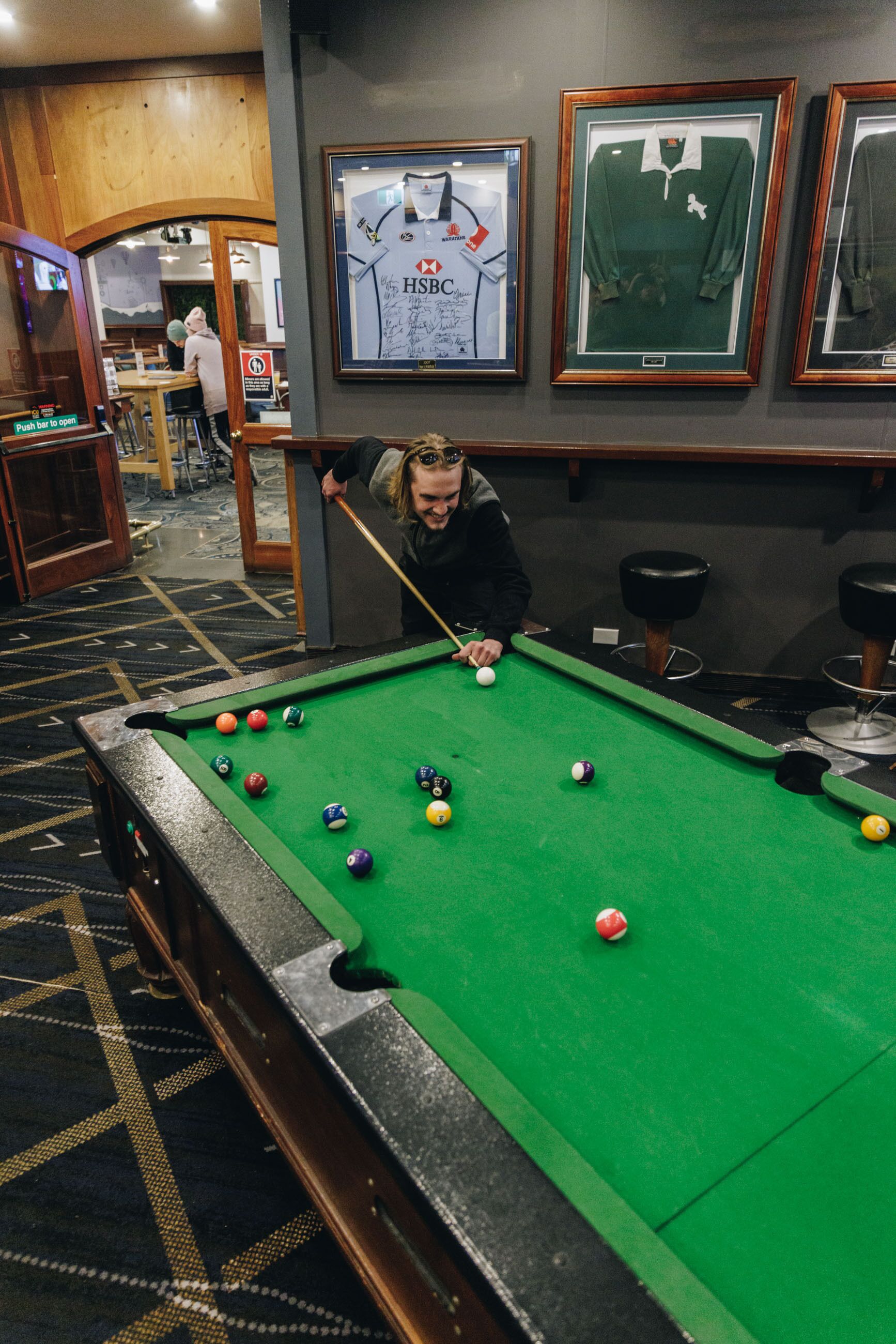 A pool table with balls, a framed jersey, and a bar area in a room with patterned carpet.