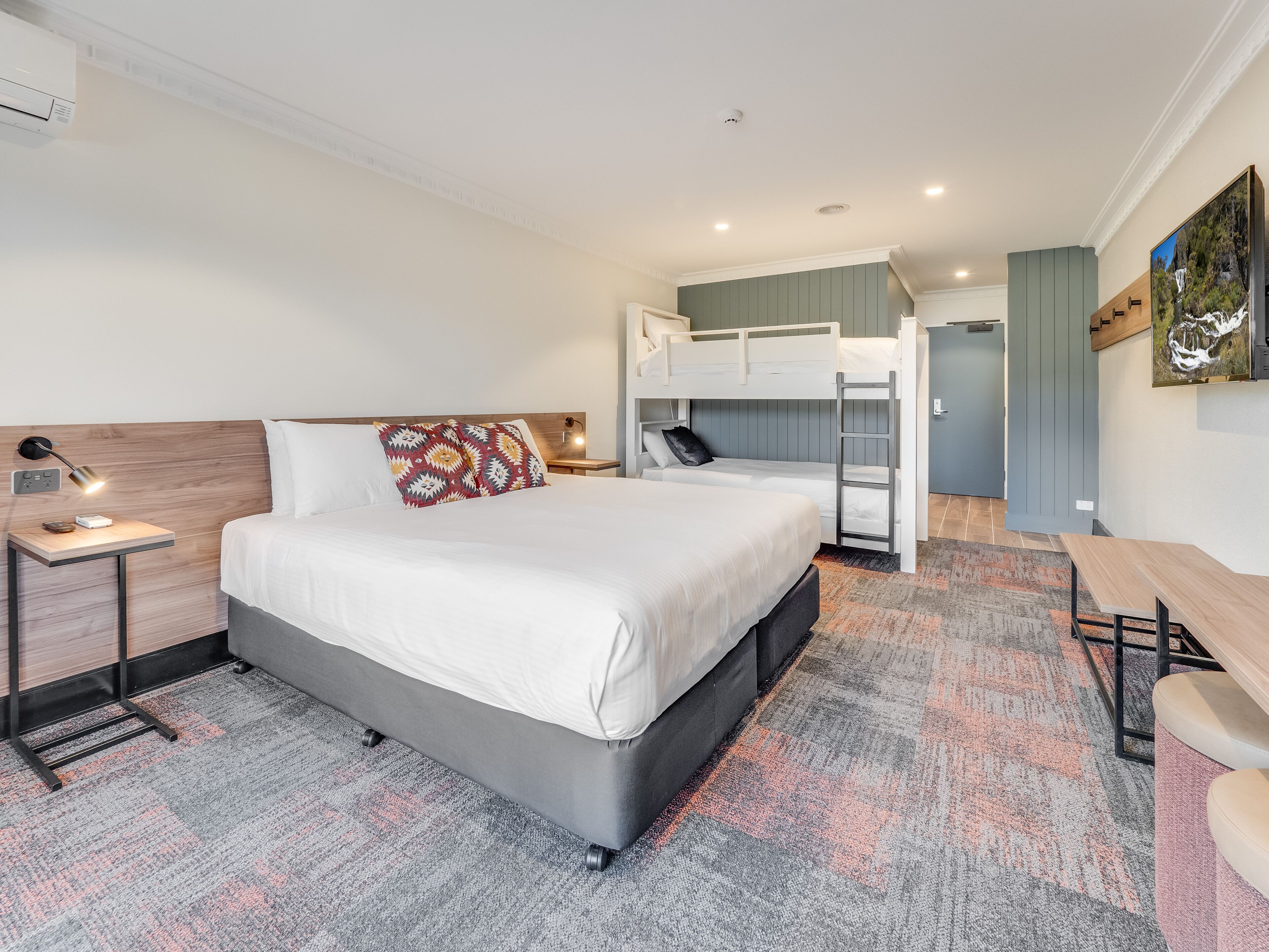 A modern hotel room with a large bed, a desk, a bunk bed, and a painting on the wall.