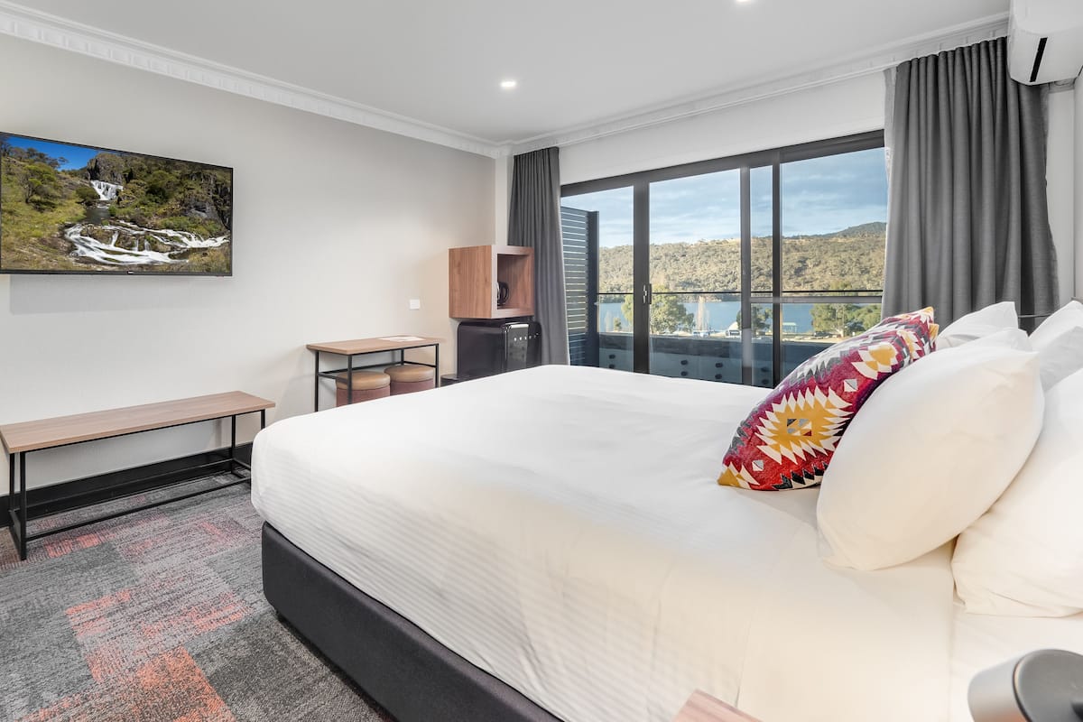 Habitación de hotel con una cama grande, un escritorio, una silla, una máquina de café y un ventanal con vistas a un lago y colinas.