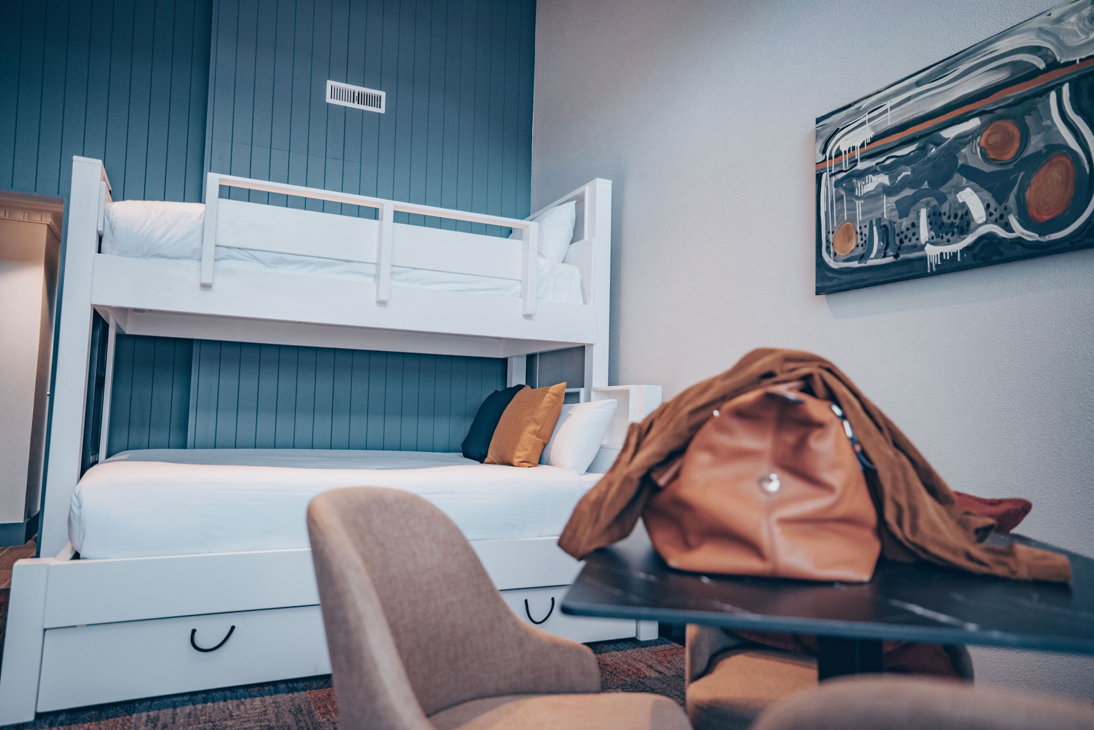 A bunk bed with a white frame and a single bed below. There is a brown leather bag on a table, and a chair is visible in the foreground.