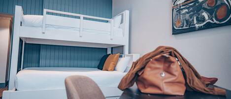 A bunk bed with a white frame and a single bed below. There is a brown leather bag on a table, and a chair is visible in the foreground.