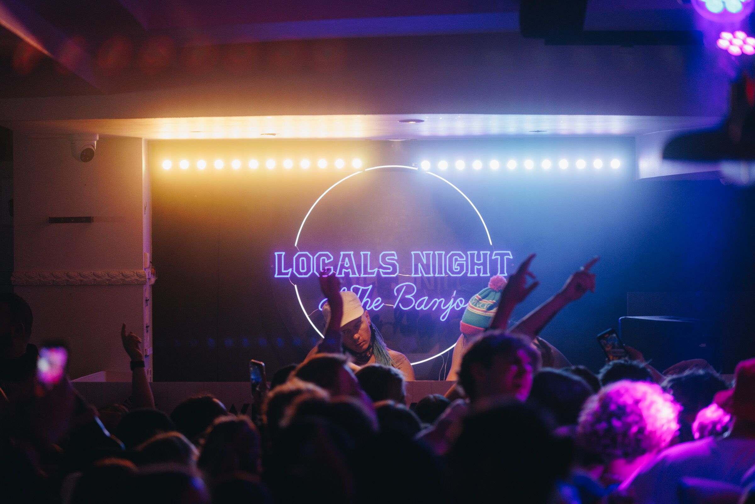 A crowded indoor event with a neon sign reading "Locals Night" and "The Banjo".