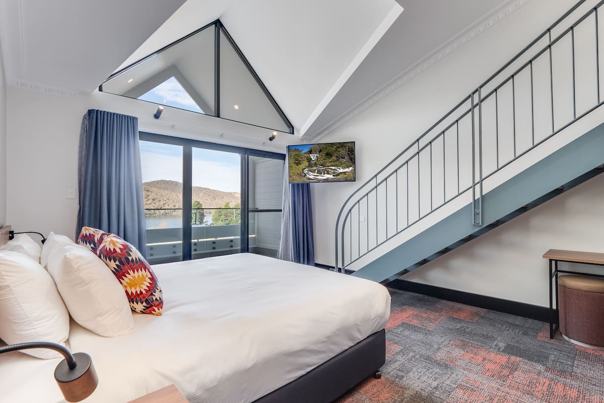 Un dormitorio con una cama grande, tragaluz, vistas al exterior, una escalera y un televisor montado.