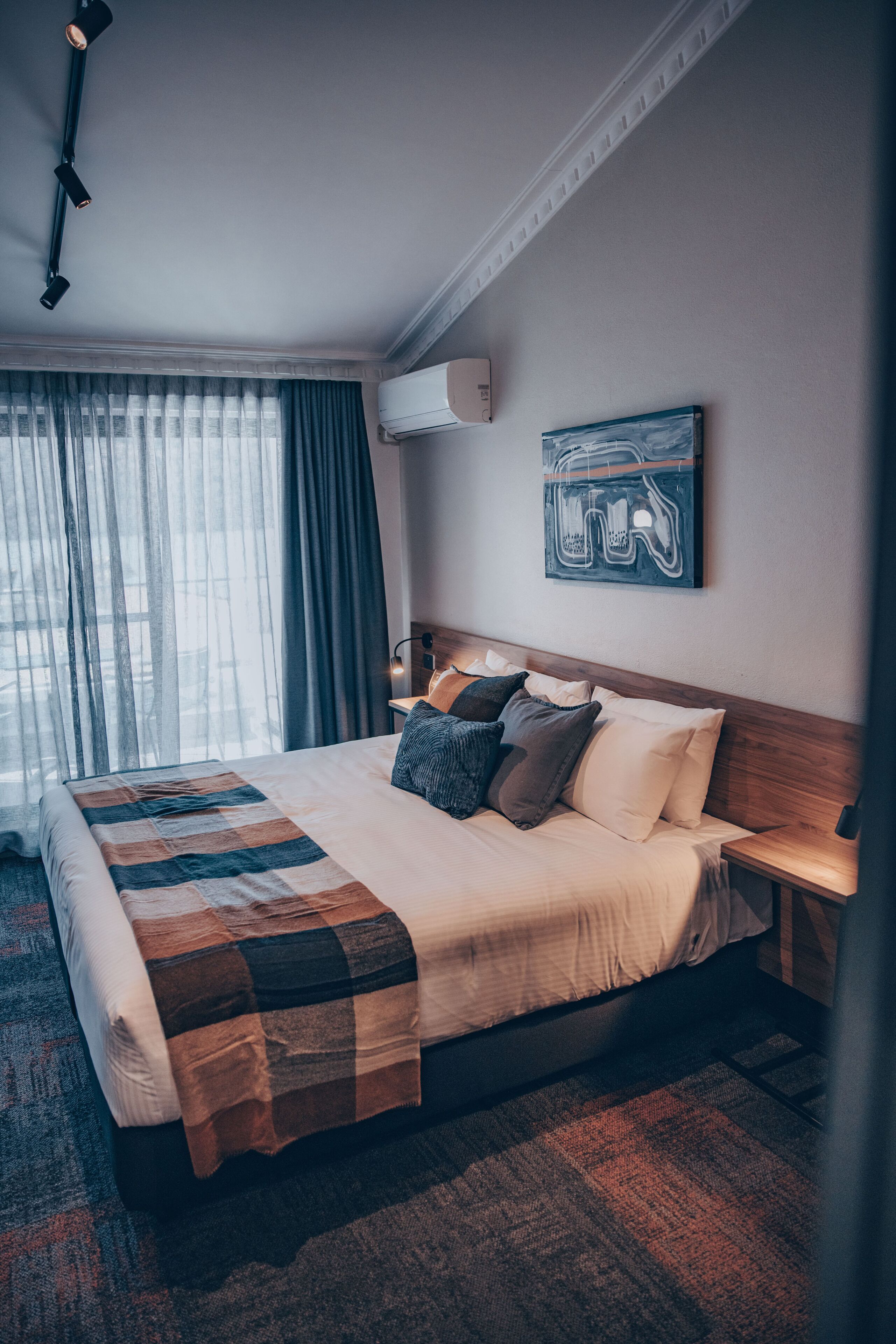 A hotel room with a large bed, a wooden headboard, a wall-mounted air conditioner, and a window with curtains.