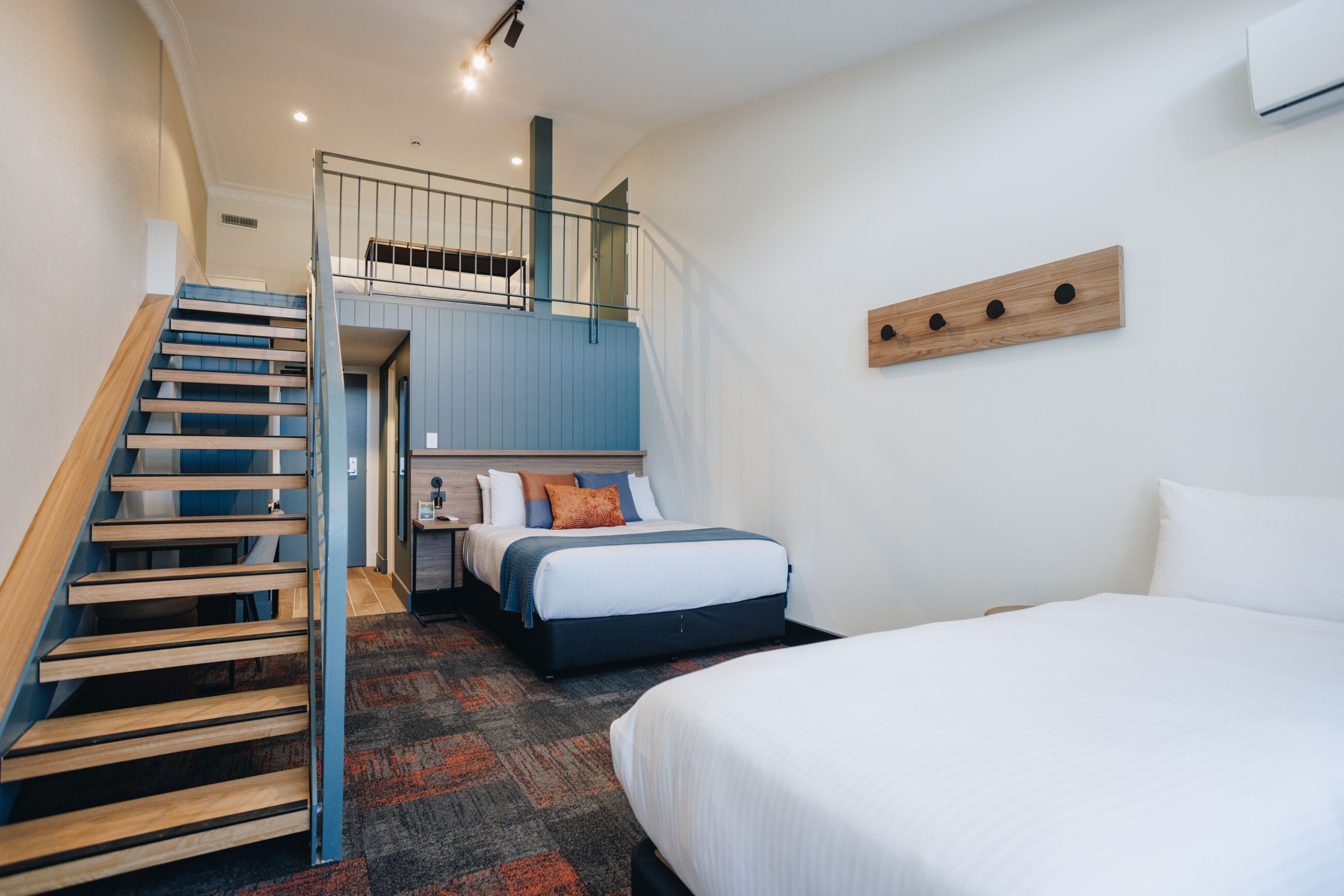 A hotel room with a wooden staircase, a bed with white bedding, a blue headboard, and a small desk with a chair.
