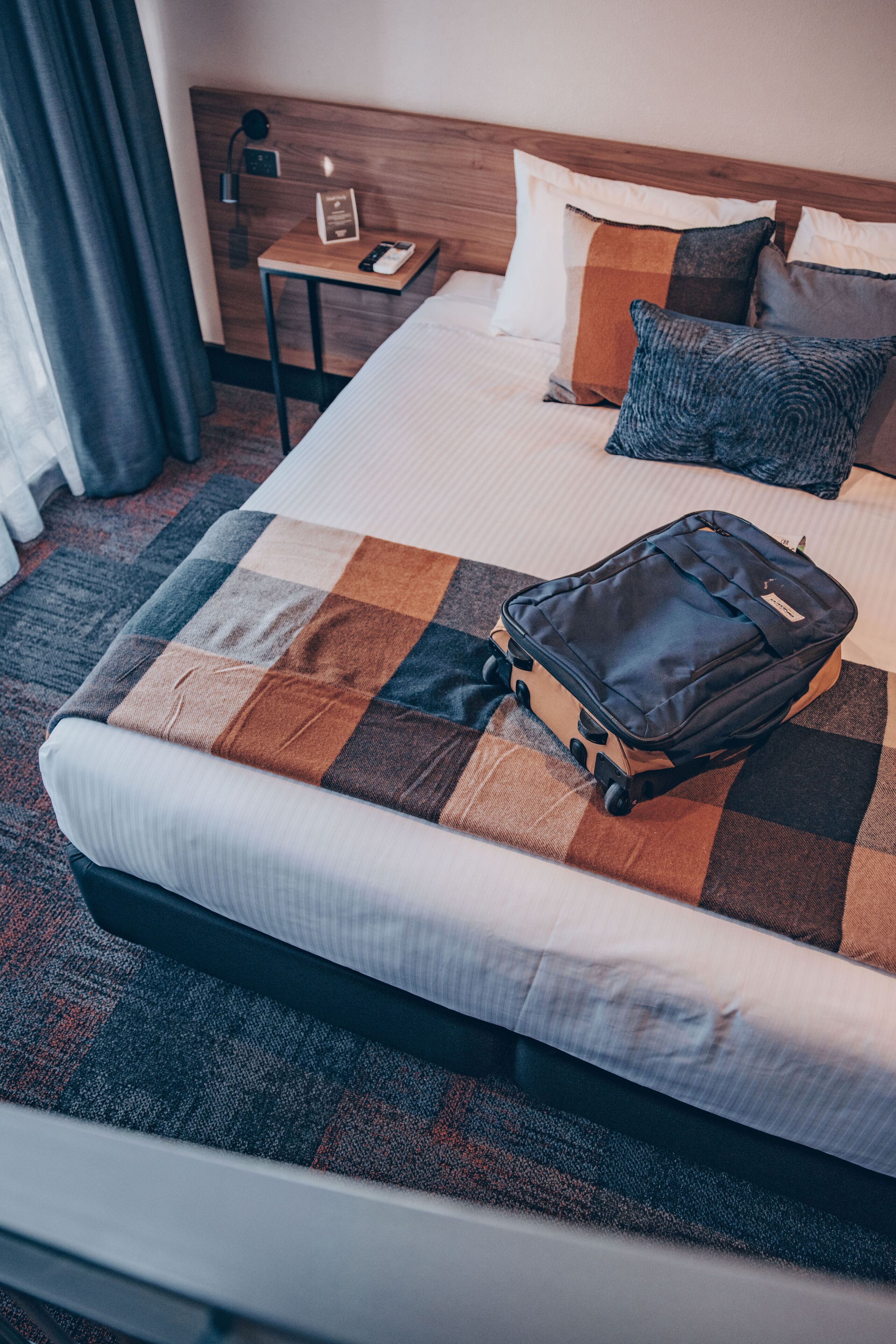 A neatly made bed with a checkered blanket, a small bedside table with a lamp, and a suitcase on the bed.