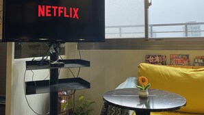 A living room with a television displaying 'NETFLIX', a small table with a flower, and a sofa with a yellow cushion. - Easymind Guesthouse, Hostel in Taipei Main Station (Taipei)