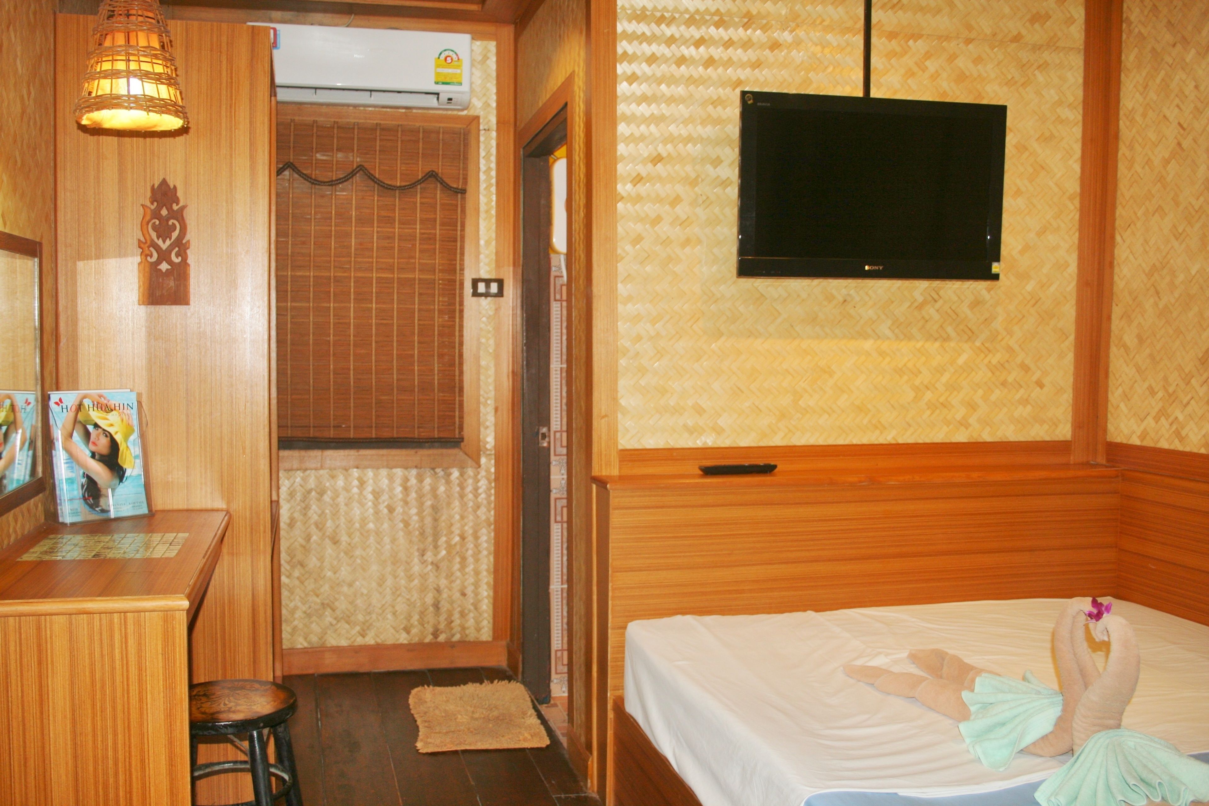 A hotel room with a wooden bed, a television mounted on the wall, a desk with a chair, and a small stool.