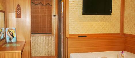 A hotel room with a wooden bed, a television mounted on the wall, a desk with a chair, and a small stool.