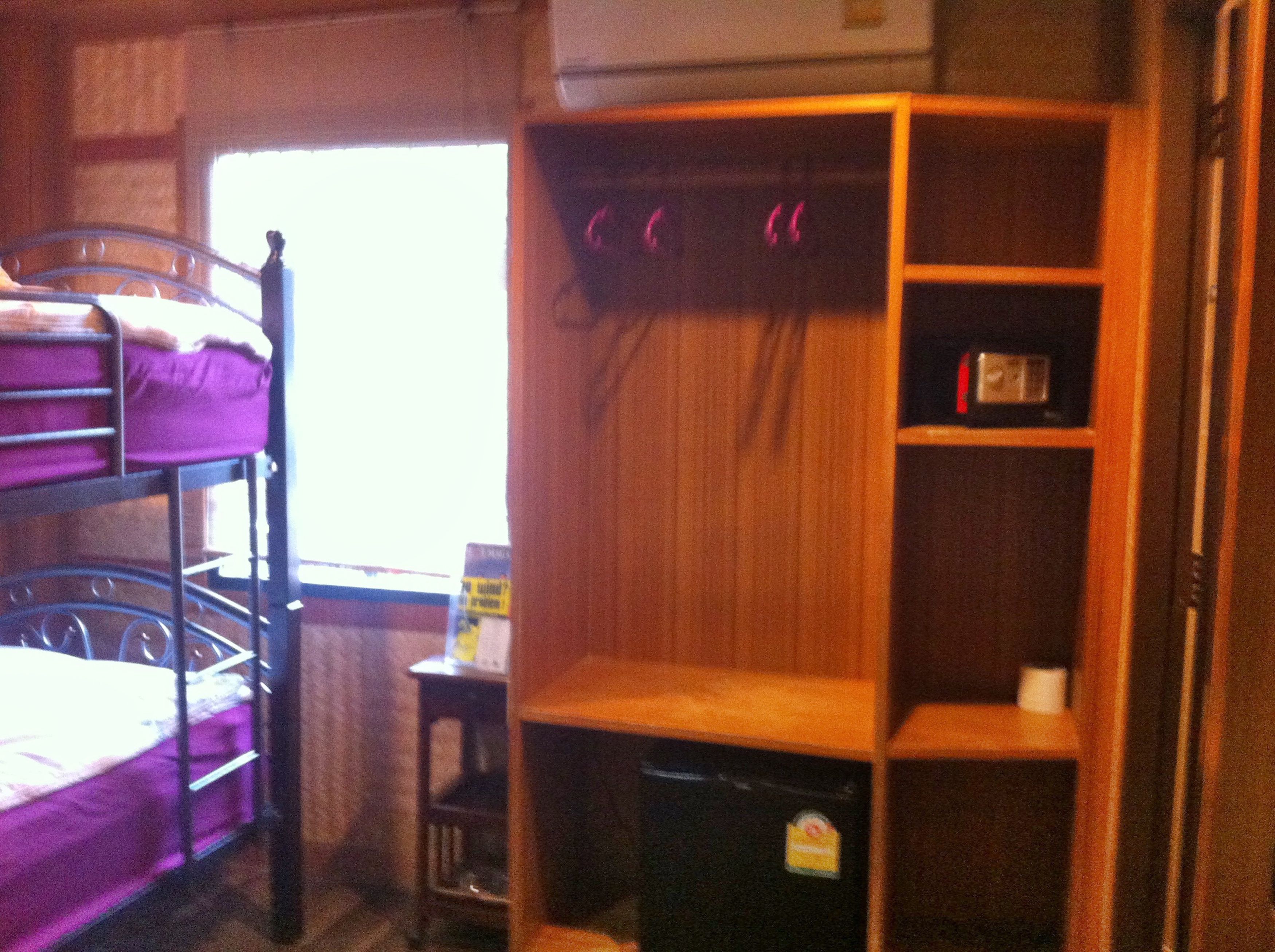 A dormitory room with bunk beds, a wooden shelf unit, a small table, and a window.