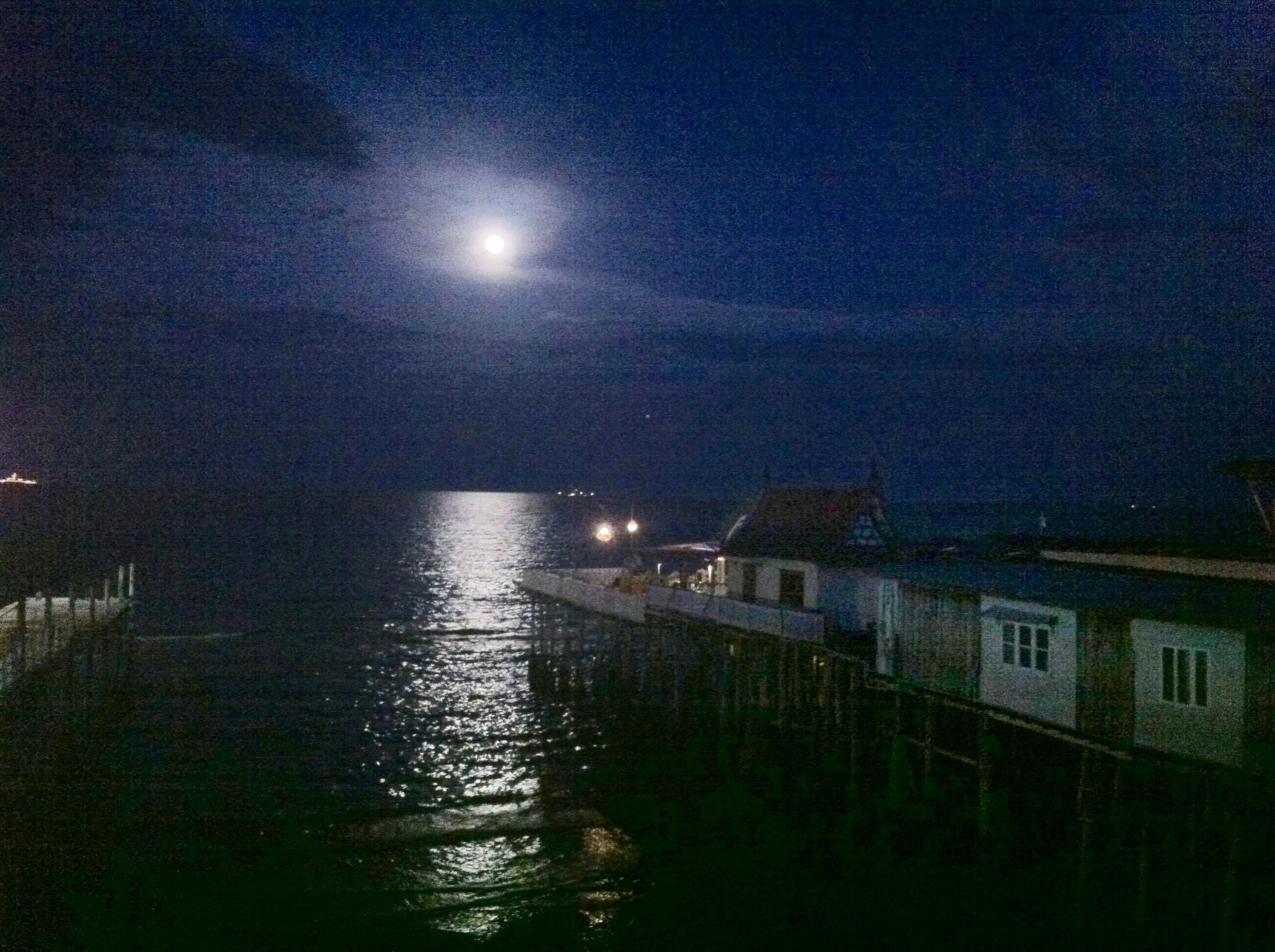 A night scene with a calm sea, a visible moon, and a pier with buildings.