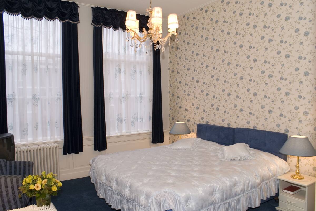 Un dormitorio con una cama grande, dos mesitas de noche con lámparas, un aparador, un radiador y una ventana con cortinas.