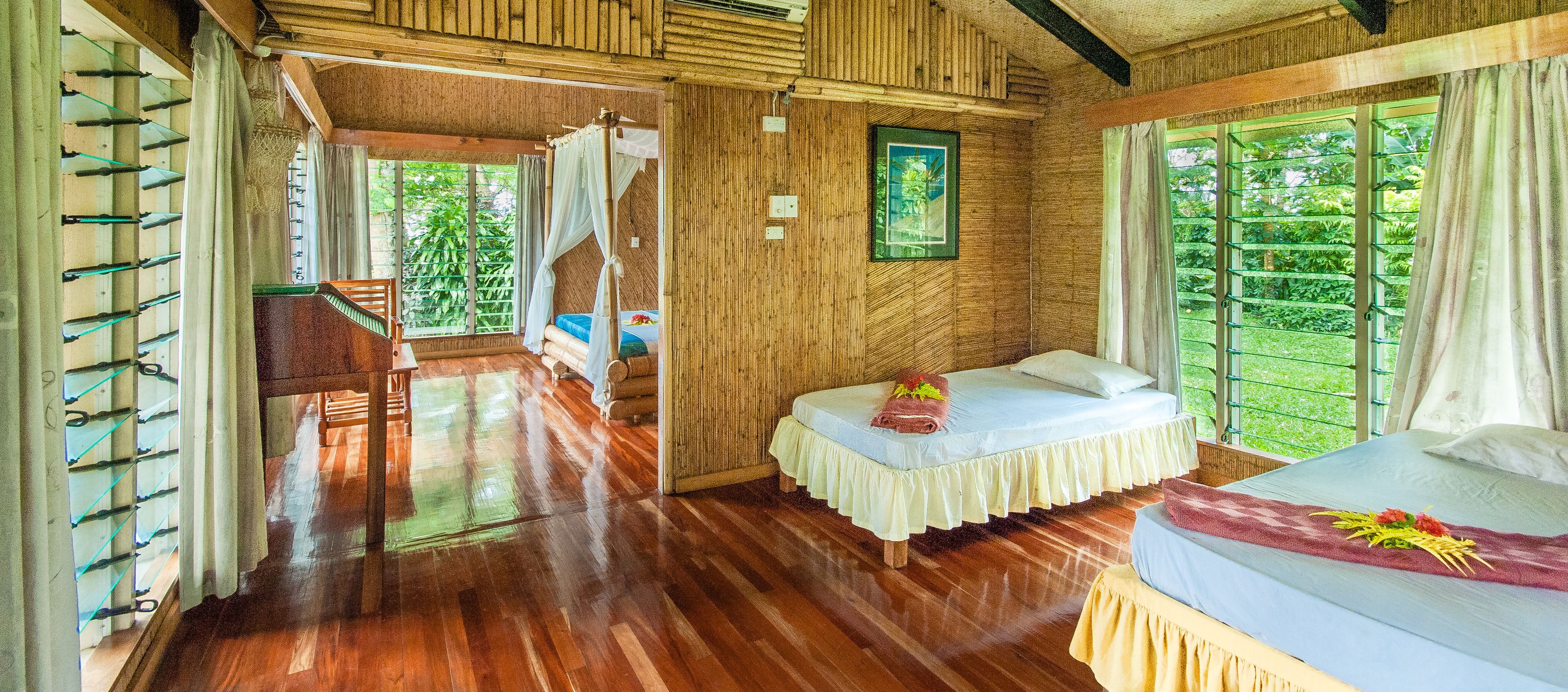 A room with wooden flooring, a bed with a white bedspread, a small table, and a window with a view of greenery.