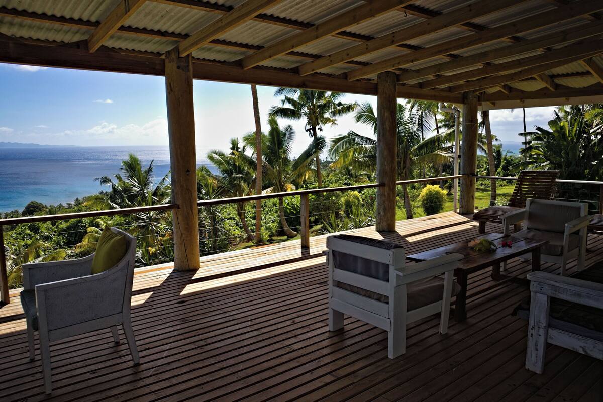 A wooden deck with outdoor furniture, overlooking a tropical landscape with palm trees and the ocean.