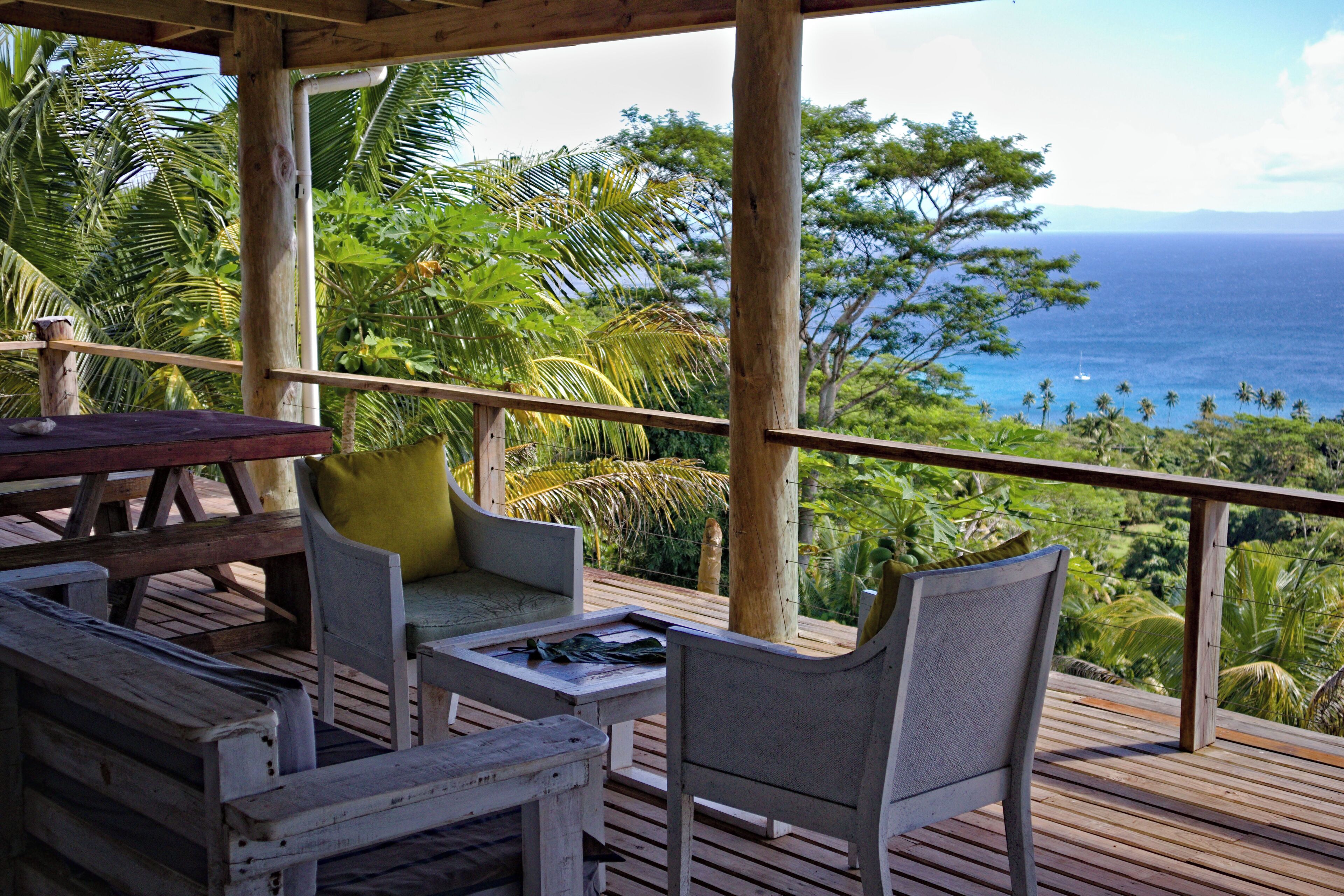 Um deck de madeira com mesa e cadeiras, com vista para uma paisagem tropical com palmeiras e o oceano.