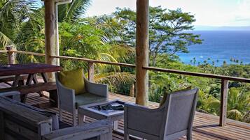 Um deck de madeira com mesa e cadeiras, com vista para uma paisagem tropical com palmeiras e o oceano.