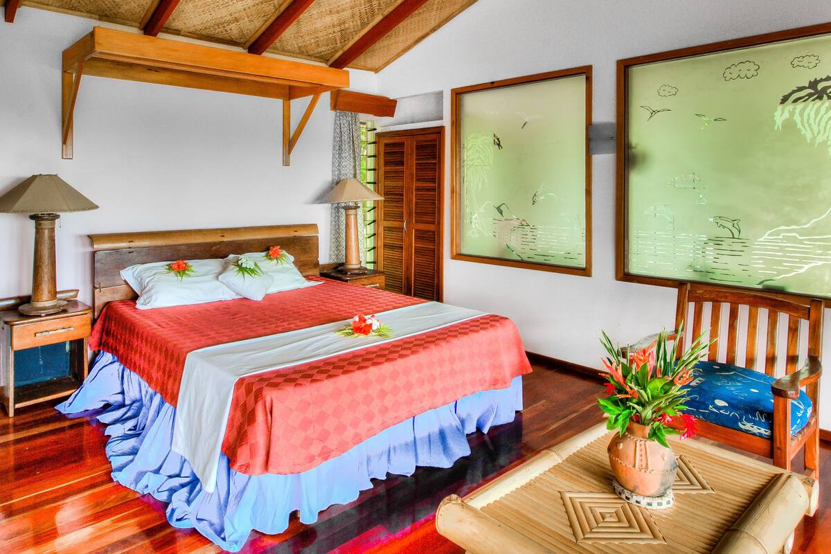 A well-lit bedroom with a wooden bed, a nightstand, a lamp, and a view of the outdoors through a window with curtains.