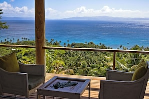 A wooden deck with two chairs and a table, overlooking a tropical landscape with palm trees and a view of the ocean.