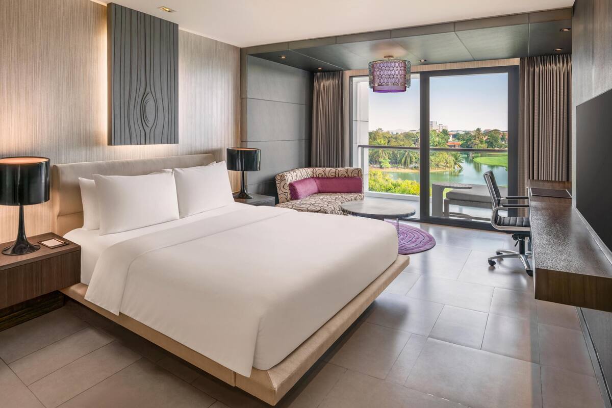 A modern hotel room with a large bed, a desk, a chair, a sofa, a TV, and a view of a golf course.