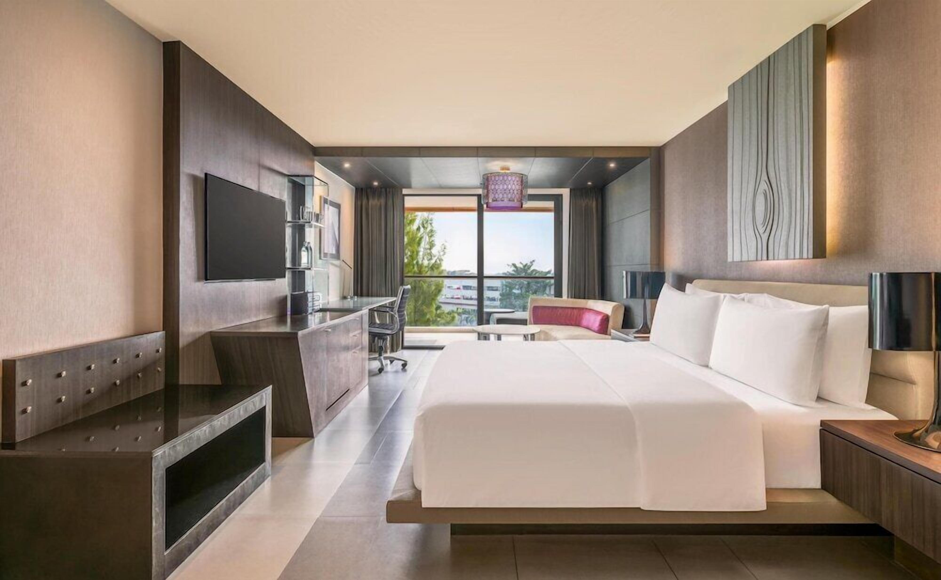 A modern hotel room with a large bed, a desk, a TV, and a balcony with a city view.