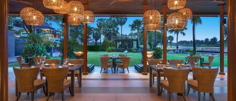 A restaurant with wicker furniture and hanging lights, overlooking a well-maintained lawn and distant golf course.