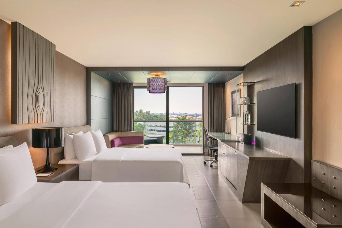 A hotel room with two beds, a desk, a TV, and a large window with a city view.